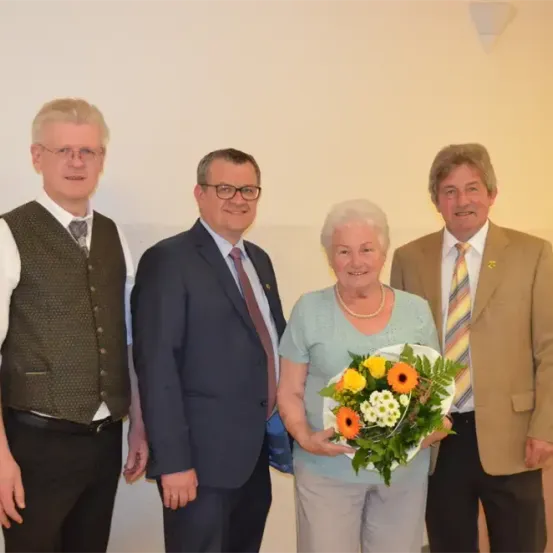 Vier Personen stehen eng beieinander in einer formellen Umgebung. Die Frau in der Mitte hält einen Blumenstrauß. Der Mann rechts trägt einen hellbraunen Anzug und eine Krawatte. Der Mann links trägt eine dunkle Weste.