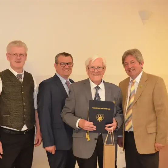 Fünf Männer stehen vor einer Wand und lächeln für ein Foto. Der Mann in der Mitte hält ein Zertifikat und eine Tasche. Die Männer auf beiden Seiten von ihm tragen Anzüge, während die anderen beiden Männer Westen tragen. Der Hintergrund ist schlicht und hell.