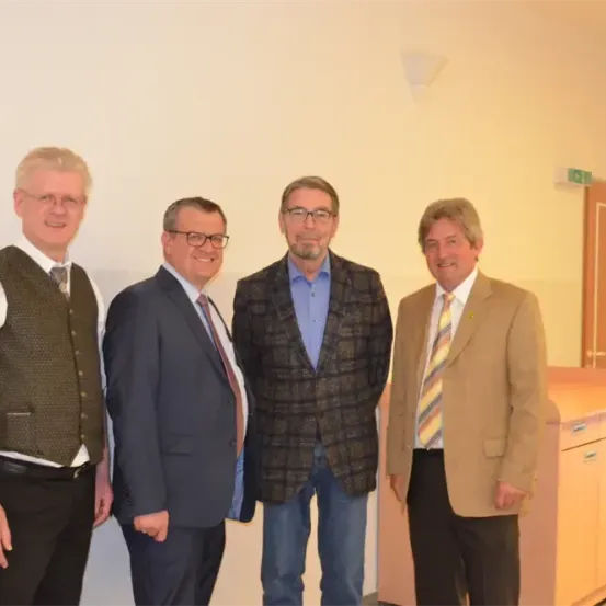 Vier Männer in formeller Kleidung stehen zusammen in einem Raum und lächeln für ein Foto. Sie tragen Brillen und Krawatten, einer trägt eine Weste. Hinter ihnen befindet sich eine weiße Wand, eine Tür und ein Holzschrank.