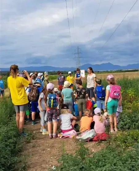 Eine Gruppe von Menschen, einschließlich Kindern, versammelt sich auf einem Feld. Einige sitzen, andere stehen, alle tragen Mützen und Rucksäcke. Der Himmel ist bewölkt.