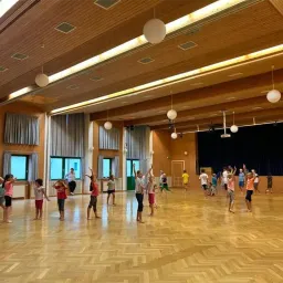 Eine Gruppe von Kindern tanzt in einem Raum mit Holzboden und Bühne. Sie sind von Fenstern mit Vorhängen und einem Projektor umgeben.