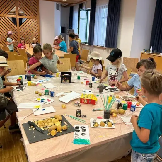 Kinder sitzen an einem Tisch in einem Klassenzimmer und malen Kartoffeln. Farben und Pinsel sind verteilt. Der Raum hat Holzwände und Stühle.
