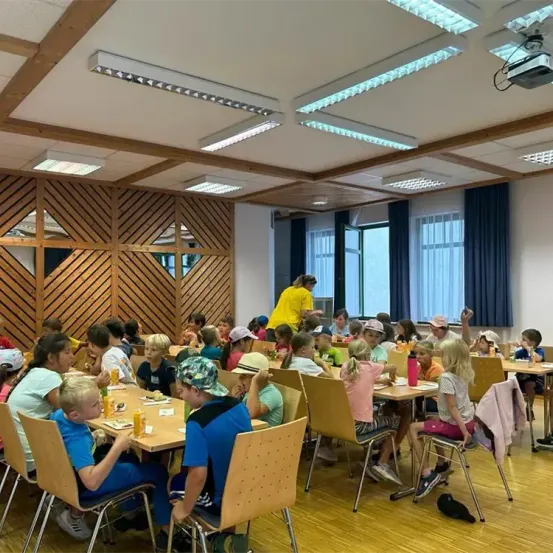 Ein Speisesaal mit Kindern an Tischen. Eine Frau in einem gelben Shirt steht hinten. Der Raum hat Holzwände und einen Projektor.