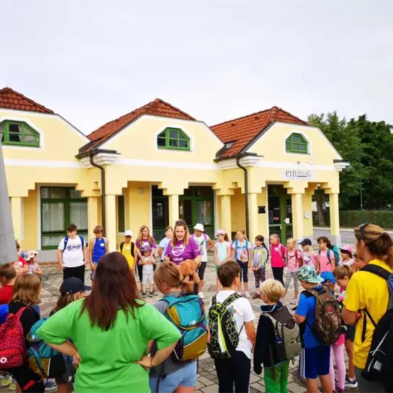 Eine Gruppe von Kindern und Erwachsenen, einige mit Rucksäcken, versammelt sich vor einem gelben Gebäude mit roten Dächern. Das Gebäude trägt ein Schild mit der Aufschrift 'Eimsbüttel'. Die Kinder schauen nach links und einige lächeln.
