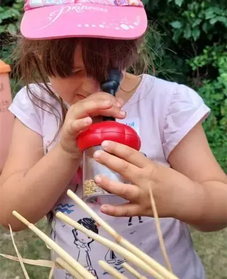 Ein kleines Mädchen mit einer rosa Prinzessin-Kappe und einem weißen T-Shirt untersucht etwas in einem roten Behälter mit schwarzem Auslass. Sie steht in einem Feld mit trockenem Gras.