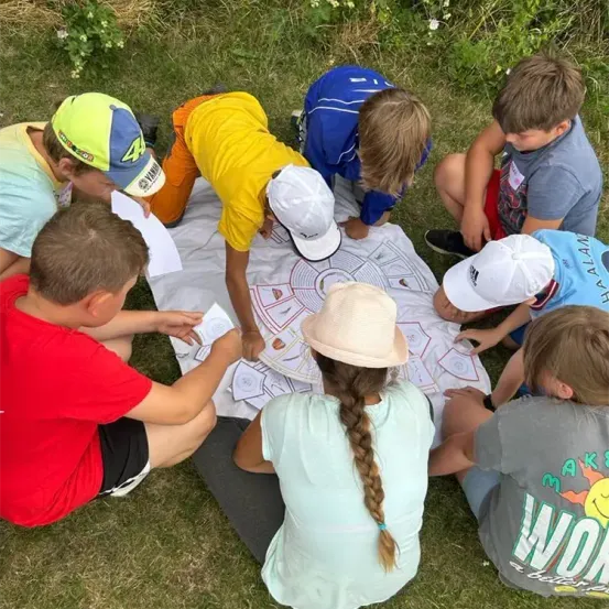 Eine Gruppe von Kindern sitzt auf dem Gras, einige stehen, um eine Karte auf einem weißen Tuch herum. Sie tragen Mützen und Turnschuhe und einige halten Papiere. Ein Mädchen hat eine geflochtene Frisur.