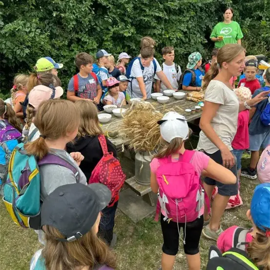 Eine Gruppe von Kindern ist um einen Tisch versammelt und lernt etwas über die Natur. Sie sind von Rucksäcken und Mützen umgeben, mit einer Frau vorne, die etwas erklärt.