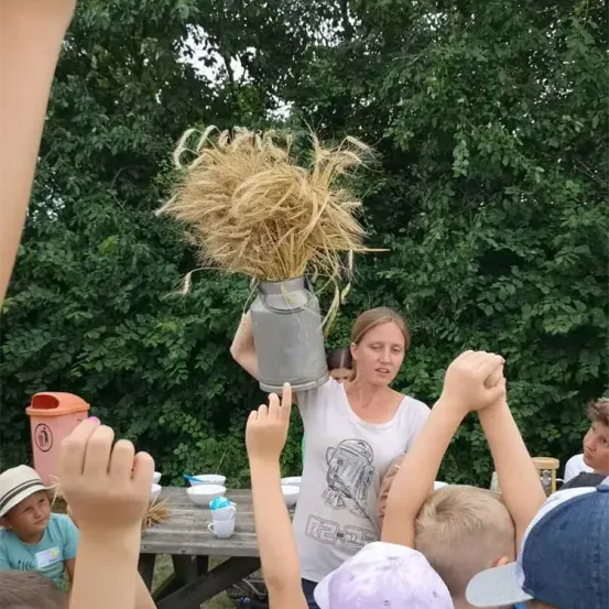 Eine Frau hält einen Eimer mit Weizen in einer Außenumgebung, umgeben von Kindern, die ihre Hände heben. Ein Tisch mit Tassen und Schüsseln ist hinter ihnen zu sehen.