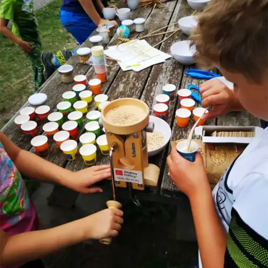 Zwei Kinder bereiten auf einem Holztisch mit Utensilien und Zutaten ein Essen zu, umgeben von Bechern und Schalen. Ein Junge rührt einen Becher mit einem Löffel um.