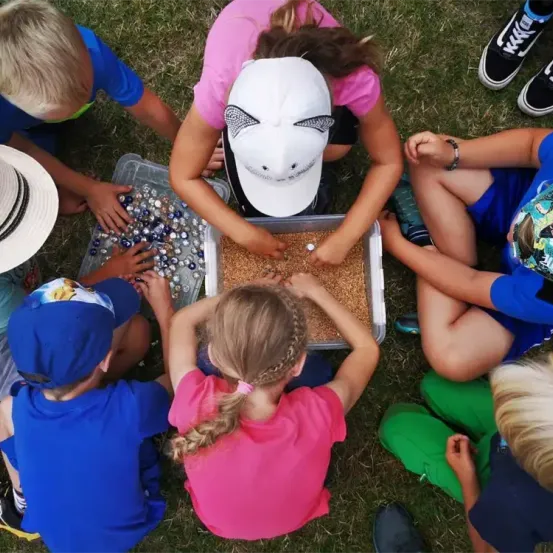 Eine Gruppe von Kindern sitzt auf dem Gras. Sie machen eine Aktivität mit einer Schale mit Perlen und Körnern. Einige von ihnen tragen Mützen und Turnschuhe.