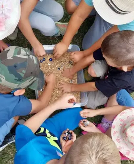 Eine Gruppe von Kindern ist um eine Schale mit kleinen Perlen und Steinen versammelt. Sie tragen Hüte und Turnschuhe. Einige von ihnen tragen Armbänder. Sie sitzen auf dem Gras.