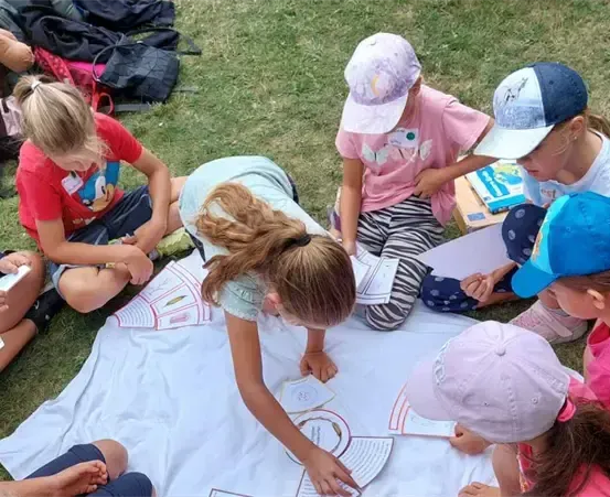 Eine Gruppe von Kindern sitzt auf einem Grasfeld, einige tragen Mützen und Turnschuhe. Sie schauen auf Papiere auf einem weißen Tuch auf dem Boden. Einige haben Rucksäcke und Bücher hinter sich.