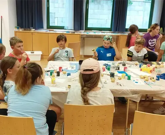 Mehrere Kinder und Erwachsene sind um einen Tisch mit verschiedenen Bastelutensilien versammelt und beschäftigen sich mit einer Bastelaktivität. Der Raum hat große Fenster und einen Holzboden.