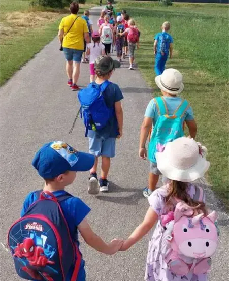 Eine Gruppe von Kindern geht Hand in Hand einen gepflasterten Weg entlang und trägt Rucksäcke. Ein Erwachsener führt sie an, und der Weg ist von grünem Gras umgeben.