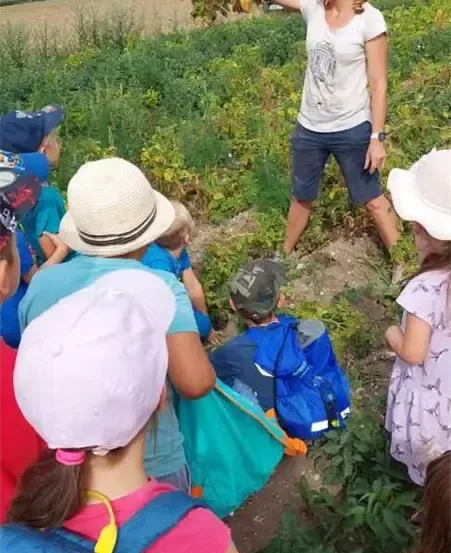 Eine Frau steht in einem Feld und hält eine Pflanze, um sie herum sitzen Kinder, alle tragen Hüte. Im Hintergrund ist ein Auto in der Nähe eines Bergrückens geparkt.