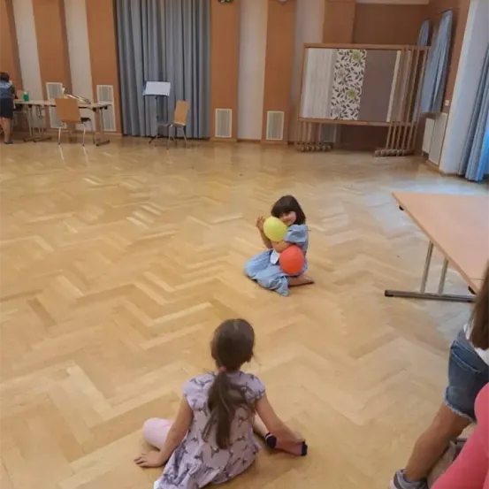 Zwei junge Mädchen spielen mit gelben und orangefarbenen Bällen in einem großen Raum mit Holzböden und Stühlen. Ein Mädchen kniet und hält die Bälle, das andere sitzt. Ein Tisch befindet sich rechts und eine Trennwand links.