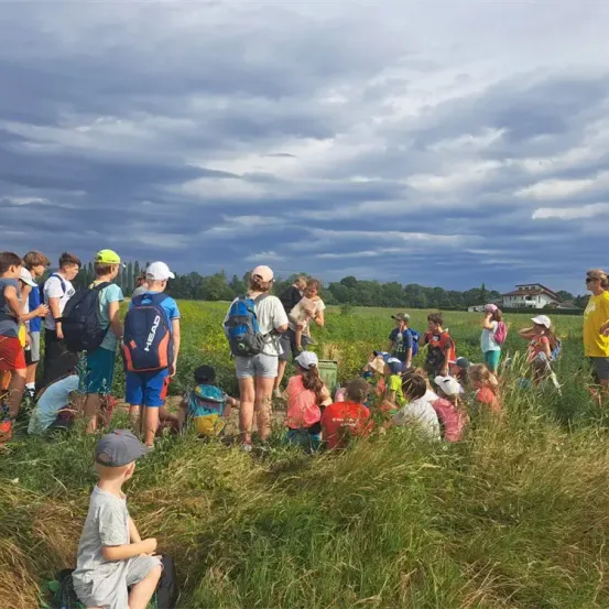 Eine Gruppe von Kindern und Erwachsenen versammelt sich auf einem Feld mit hohem Gras, einige sitzen und andere stehen. Sie tragen Hüte und haben Rucksäcke dabei. Der Himmel ist bewölkt.
