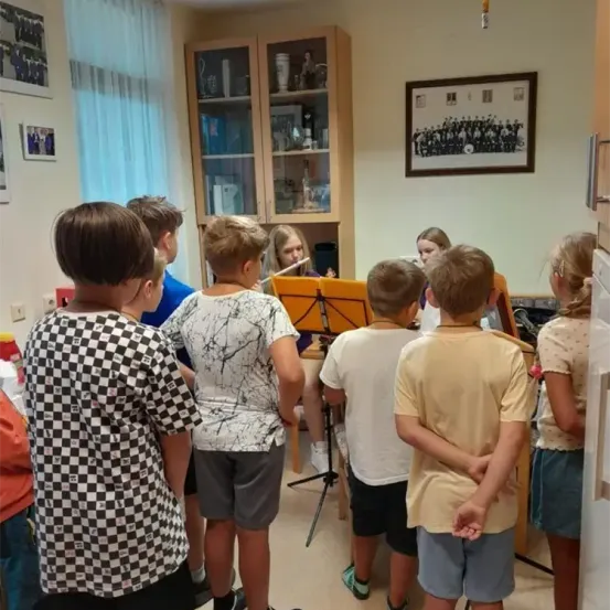 In einem Raum stehen Kinder um eine Frau herum, die Flöte spielt, mit einem Schrank und einem gerahmten Bild im Hintergrund.