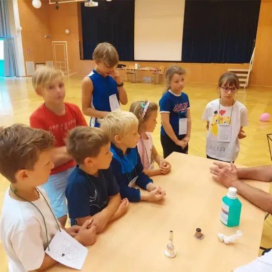Eine Gruppe von Kindern nimmt an einem wissenschaftlichen Experiment in einem Raum mit Holzböden und Vorhängen teil. Sie stehen um einen Tisch mit einer Flasche, einer Glocke und Taschentüchern.