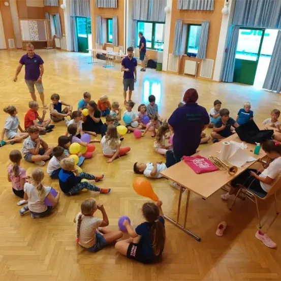 Kinder sitzen im Kreis und halten Luftballons, während zwei Erwachsene in der Nähe stehen. Ein Erwachsener hält eine Trompete und ein rotes Tuch. Ein Tisch hat eine Papierrolle und eine Flasche. Der Raum hat Holzböden und blaue Vorhänge.
