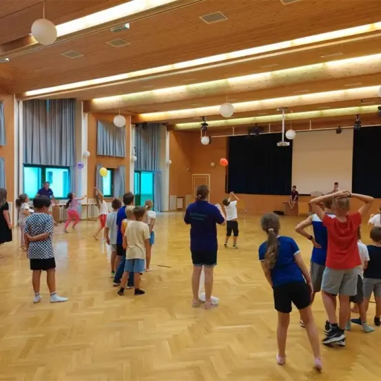 Eine Gruppe von Kindern und Erwachsenen befindet sich in einer Turnhalle. Einige Kinder spielen mit Bällen, während andere zuschauen. Die Turnhalle hat einen Holzboden, weiße Wände und eine Projektionsleinwand.