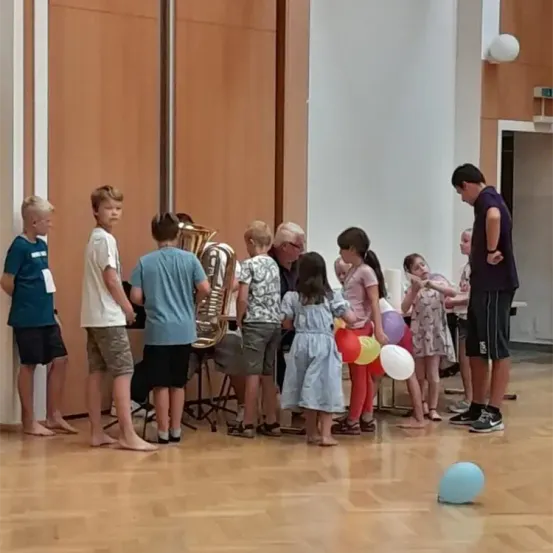 Eine Gruppe von Kindern und Erwachsenen befindet sich in einem Raum und beobachtet einen Mann, der Tuba spielt. Die Kinder halten Luftballons und schauen neugierig zu.