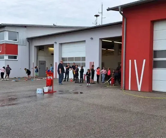 Mehrere Menschen stehen vor einer Feuerwehrwache, wobei zwei Personen den Boden vor ihr reinigen. Das Gebäude hat große weiße Garagentore.