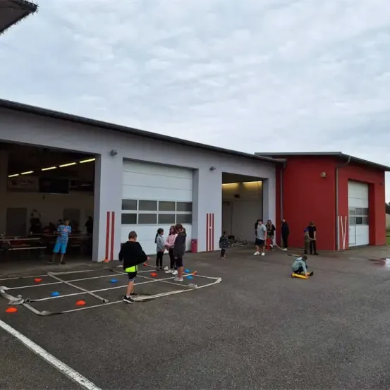 Mehrere Menschen versammeln sich vor einem großen Gebäude mit Garagentoren und sind in einem Spiel auf einem Gitterfeld mit orangefarbenen und blauen Kegeln engagiert. Das Wetter ist bewölkt.