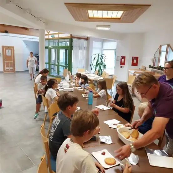 Eine Gruppe von Kindern sitzt um einen langen Tisch in einem Speisesaal, während ein erwachsener Mann Essen serviert. Eine Frau geht im Hintergrund.