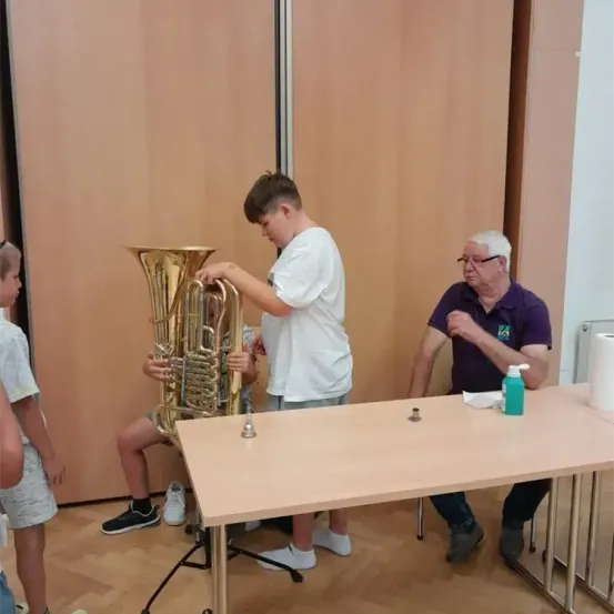 Ein junger Junge hält eine große Messingtuba, neben ihm sitzt ein älterer Mann. Sie sind in einem Raum mit Holzwänden und -boden. Ein weiterer Junge sitzt auf dem Boden.