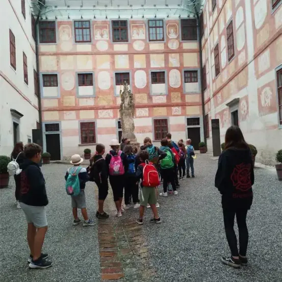 Eine Gruppe von Kindern mit Rucksäcken steht im Innenhof eines historischen Gebäudes. Der Innenhof ist von Gebäuden mit dekorativen Elementen umgeben. In der Mitte befindet sich eine Statue.