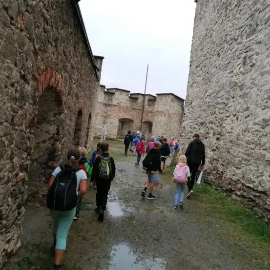 Eine Gruppe von Kindern mit Rucksäcken geht auf einem Pfad zwischen zwei Steinmauern einer alten Burg. Einige tragen Turnschuhe, andere sind barfuß.