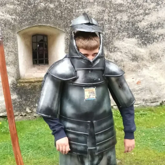 Ein junger Junge in einer mittelalterlichen Ritterrüstung steht neben einer Steinmauer mit einem vergitterten Fenster. Er steht mit dem Rücken zur Kamera.