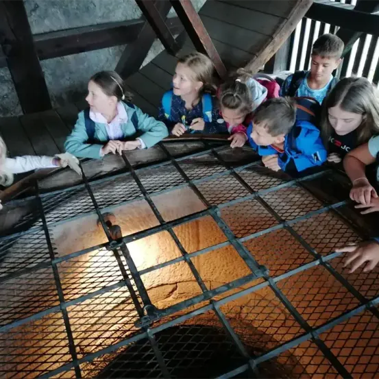 Mehrere Kinder sehen durch ein Metallgitter in eine Grube, wahrscheinlich auf einer Klassenfahrt. Sie tragen Rucksäcke und Jacken, und eine Holztreppe ist hinter ihnen.