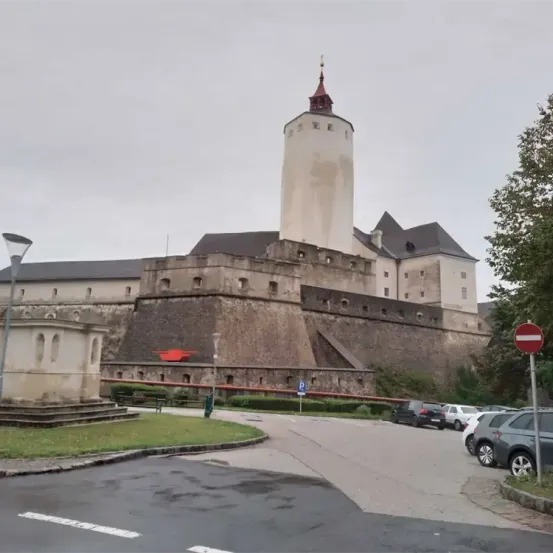 Eine große mittelalterliche Burg mit einem hohen Turm steht gegen einen bewölkten Himmel. Die Burg hat ein rotes Dach und viele Fenster. Vor der Burg befindet sich ein kleiner Garten und ein rotes und weißes Schild.