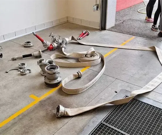 Verschiedene Feuerwehrschläuche und Teile liegen auf einem Betonboden. Drei Personen stehen im Hintergrund.