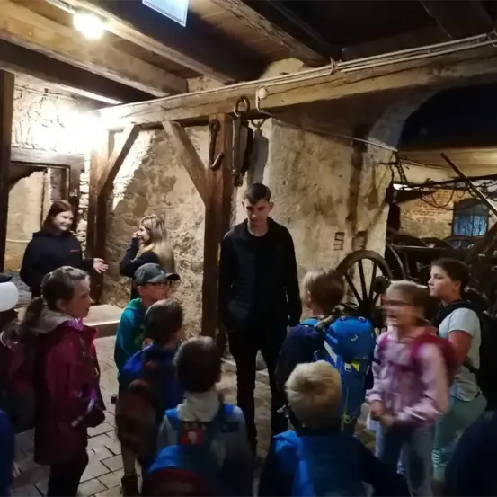 Eine Gruppe von Kindern und Erwachsenen befindet sich in einer historischen Umgebung, wahrscheinlich einem Museum. Die Kinder tragen Rucksäcke und scheinen einem Mann in einem schwarzen Outfit zuzuhören.