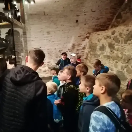 Eine Gruppe von Kindern versammelt sich in einem Raum mit einer Steinmauer. Ein Erwachsener erklärt ihnen etwas. Einige Kinder schauen nach oben.