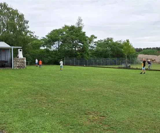 Mehrere Personen spielen Fußball auf einem Rasenfeld mit einer Hütte und einem Zaun im Hintergrund.