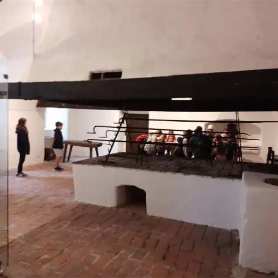 Ein Raum mit gefliesten Böden, weißen Wänden und einer Holzbank. Mehrere Personen sitzen auf dem Boden, während andere in der Nähe stehen.