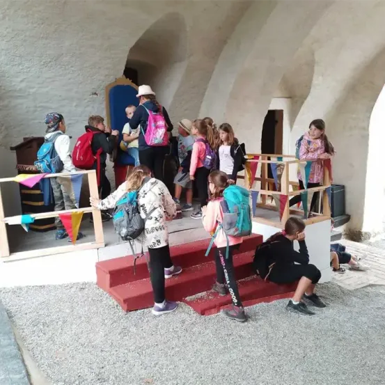 Eine Gruppe von Kindern, einige mit Rucksäcken, versammelt sich in der Nähe eines Throns. Sie scheinen sich auf eine Aktivität oder Veranstaltung vorzubereiten. Der Bereich ist von Wänden mit Bögen umgeben und beinhaltet eine rote Treppe.