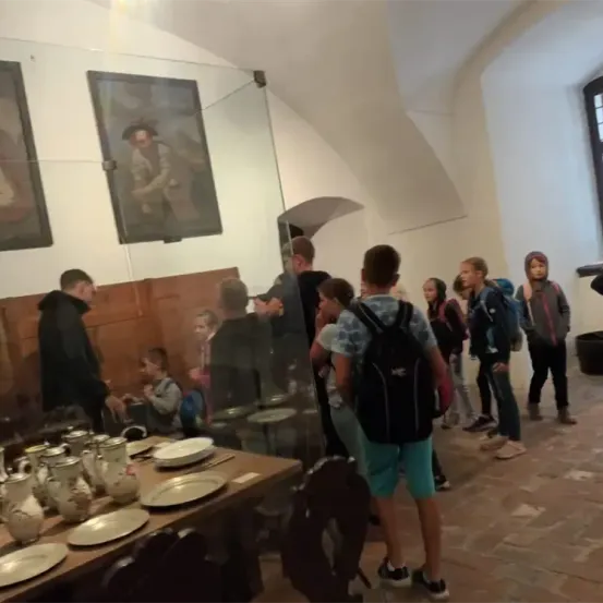 Eine Gruppe von Menschen, wahrscheinlich Schüler, steht in einem Raum mit einem Holztisch und Stühlen. Zwei Gemälde hängen an der Wand, und ein Mann mit einem Rucksack steht vor einer Glaswand.