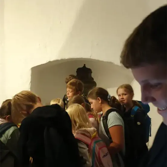 Eine Gruppe von Kindern mit Rucksäcken steht in einem Raum, einige schauen nach unten, andere seitlich.