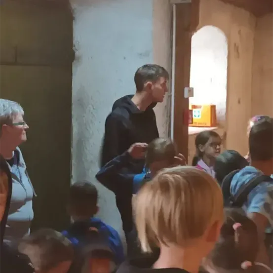 Eine Gruppe von Kindern und Erwachsenen befindet sich in einem Raum. Ein Mann in Schwarz steht in der Nähe der Tür, und eine Frau mit Brille steht links. Der Raum hat eine gelbe Box an der Wand.
