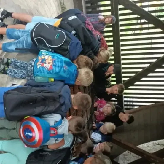 Eine Gruppe von Kindern ist um eine Holzkonstruktion versammelt, einige tragen Rucksäcke und Turnschuhe. Sie schauen nach unten, möglicherweise auf etwas Interessantes am Boden.