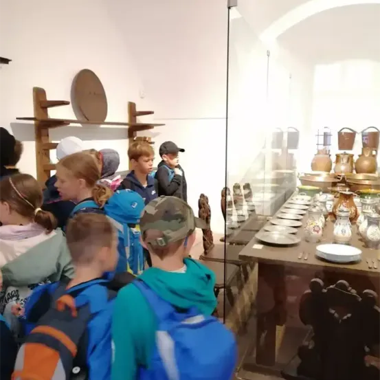 Eine Gruppe von Kindern mit Rucksäcken versammelt sich vor einer Glasvitrine und betrachtet einen gedeckten Tisch mit Tellern, Besteck und dekorativen Vasen darin.