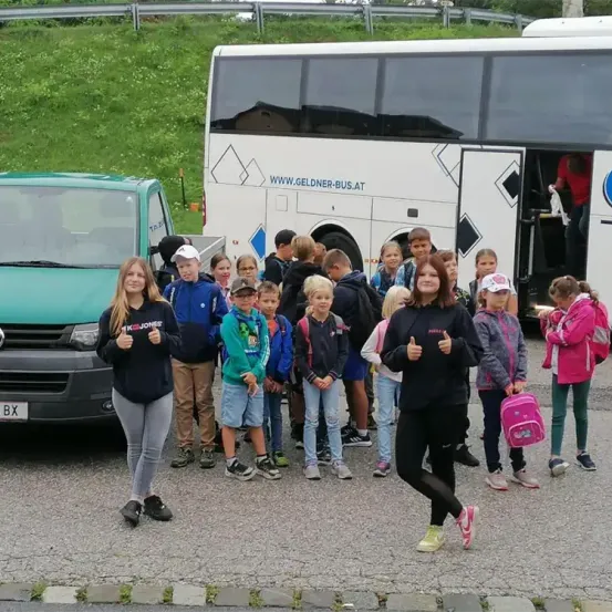 Eine Gruppe von Kindern steht neben einem Bus und einem Lkw, eine Frau gibt Daumen hoch. Im Hintergrund befindet sich ein Grashügel und ein Zaun.