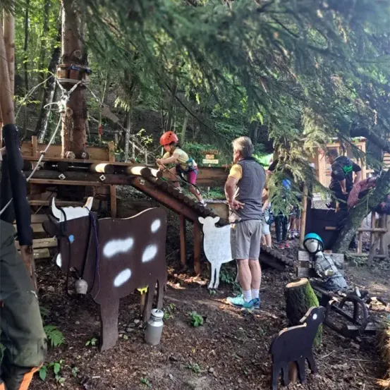 Ein Kind mit rotem Helm klettert auf eine Holzkonstruktion im Wald. Ein erwachsener Mann beobachtet. In der Nähe befinden sich eine Kuhstatue, eine Milchkanne und eine Schafsstatue. Bäume und Holzkonstruktionen umgeben das Gebiet.