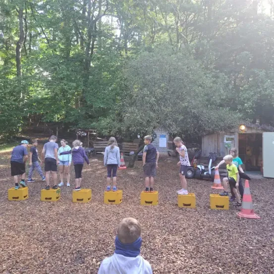 Eine Gruppe von Kindern steht auf gelben Kisten in einem bewaldeten Bereich, wahrscheinlich für ein Spiel. Hinter ihnen befinden sich Verkehrskegel und ein Gebäude.