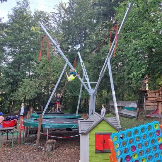 Zwei Kinder schwingen auf Schaukeln über einem Trampolin. Eine Frau beobachtet von der Seite. Ein Haus, Spielzeug und eine Rutsche sind im Hintergrund.